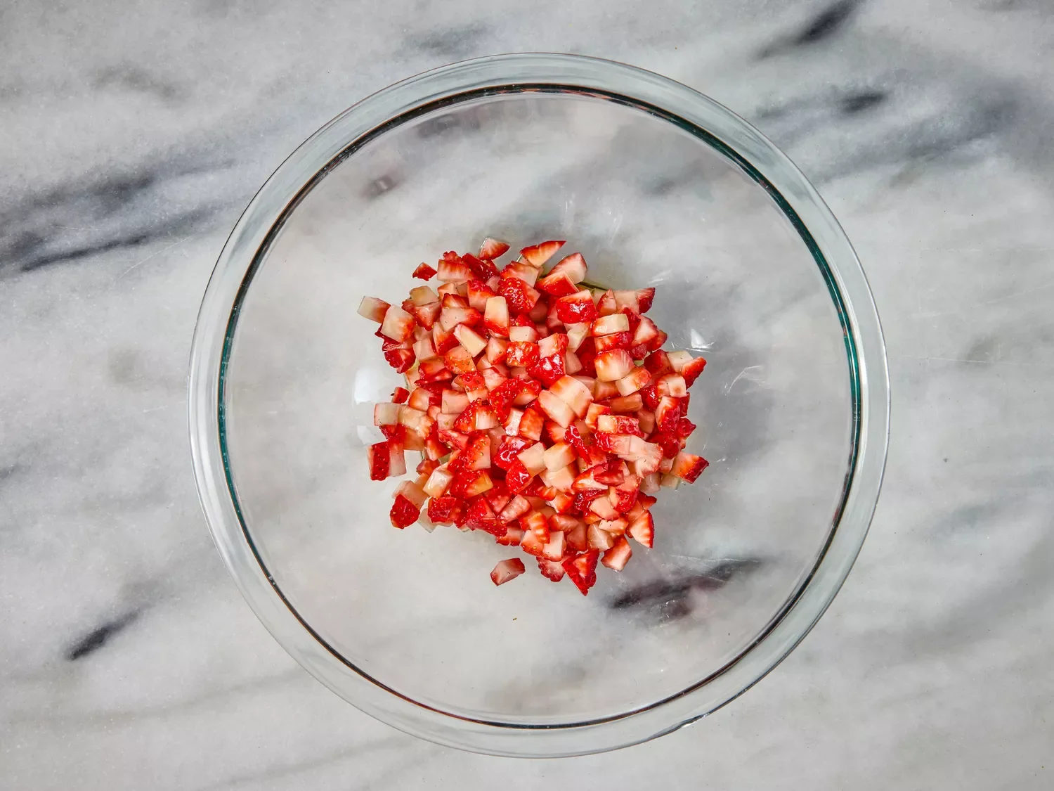 Chopped fresh strawberries in a glass bowl on a marble countertop.